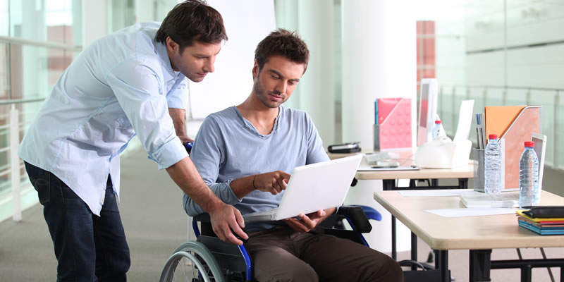 A man in a wheelchair speaks to a coworker at his desk