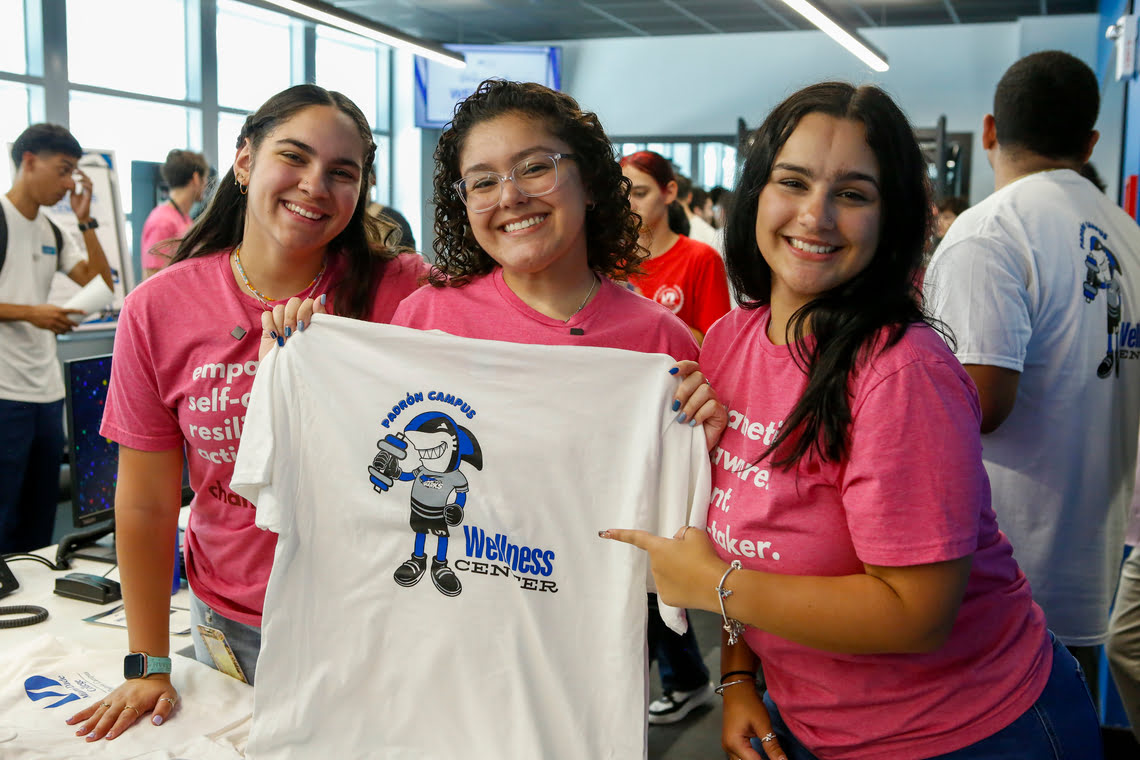 Three smiling young women holding a Padrón Campus Wellness Center t-shirt.