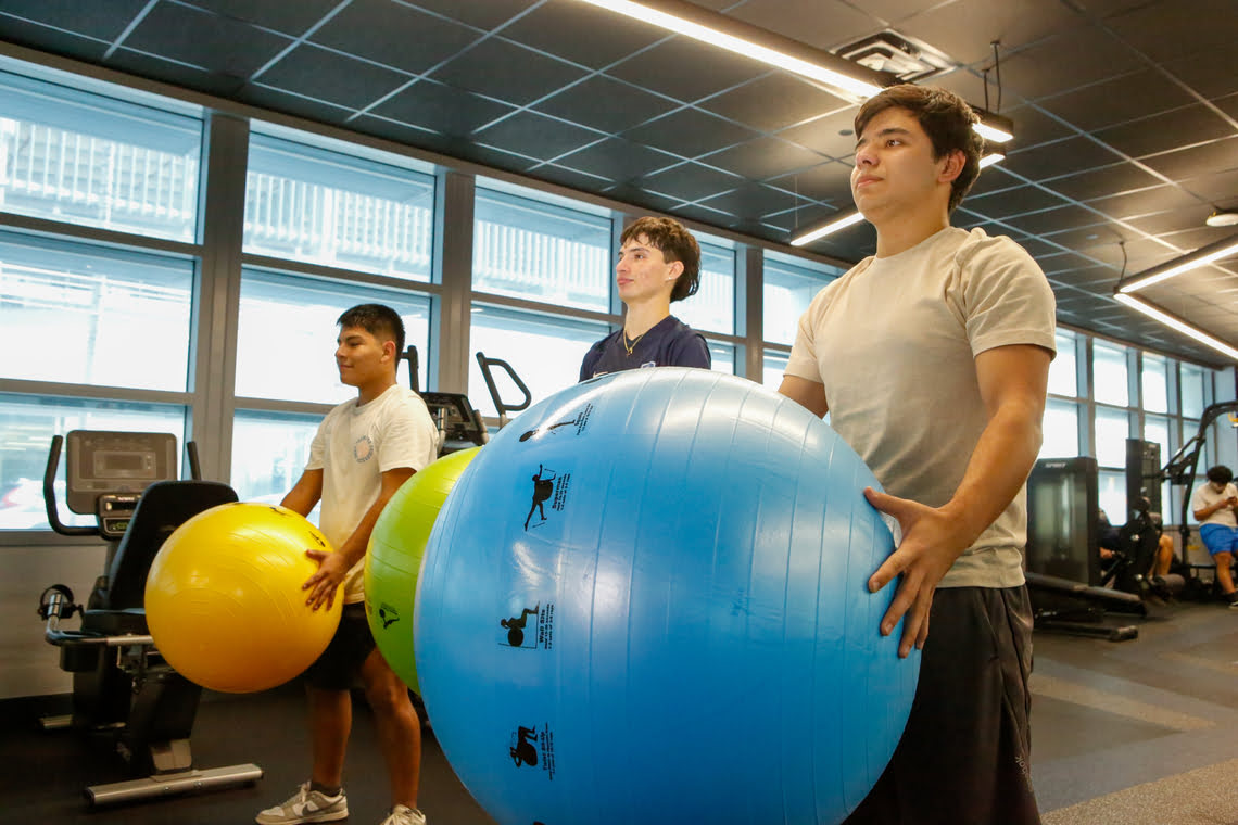 Three young men in a gym holding large exercise balls during a fitness class.