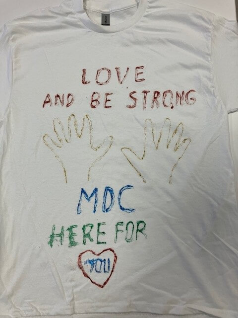 white tee shirt with the words 'love and be strong; MDC here for you' hand painted on it