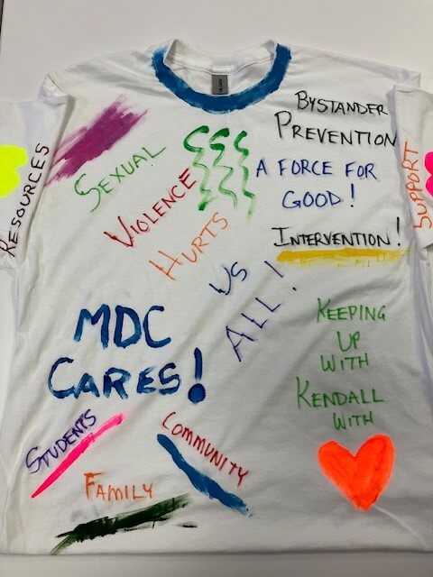 white tee shirt with the words 'sexual violence hurts us all! MDC cares! Intervention! Bystander prevention! A force for good! Keeping up with Kendall with love' hand painted on it